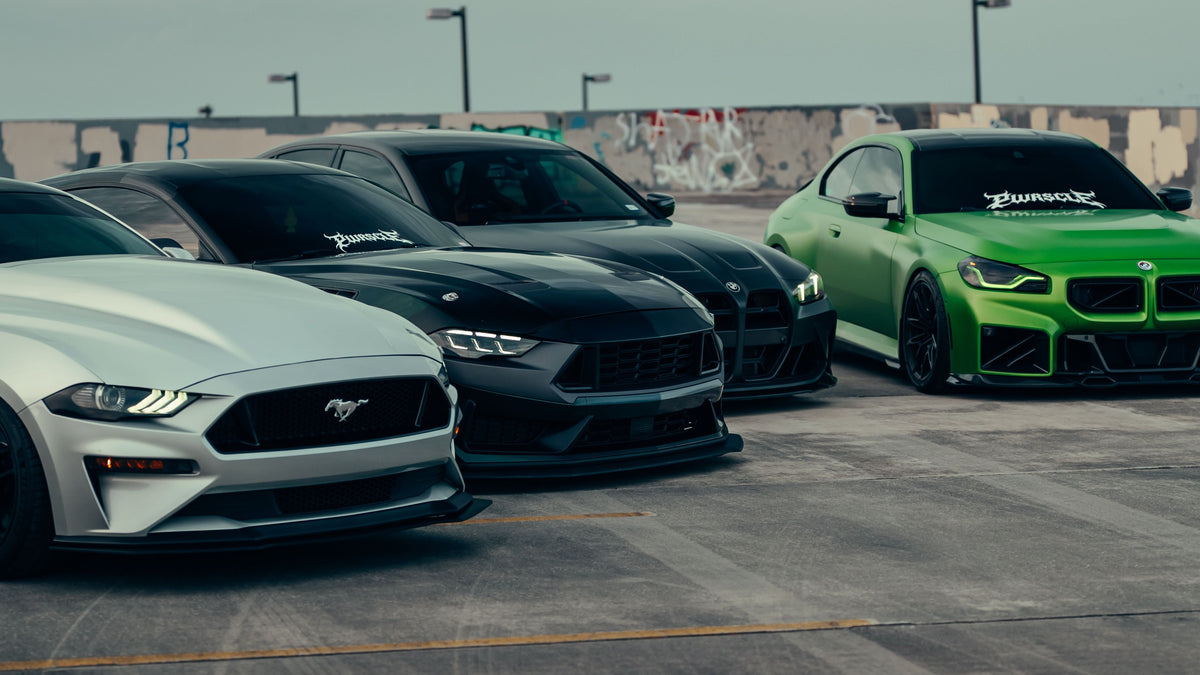 Three high-performance cars parked outdoors on a racetrack: a silver Ford Mustang in the foreground, a black Ford Mustang behind it, and a bright green BMW M4 on the right. The BMW M4 has a visible BurnerX decal on the windshield.