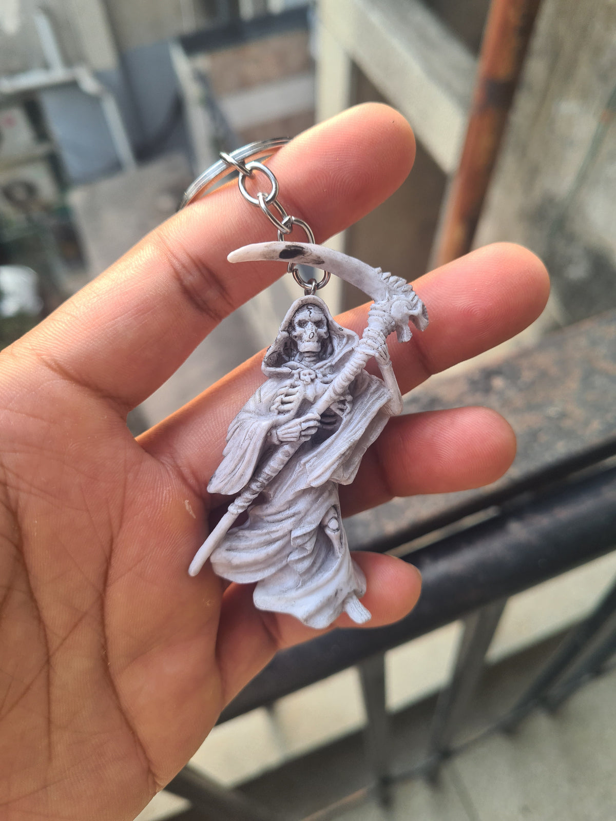 A hand holding a small gray plastic keychain featuring a grim reaper figurine with a large horned skull and a silver metal key ring, set against a blurred outdoor background.