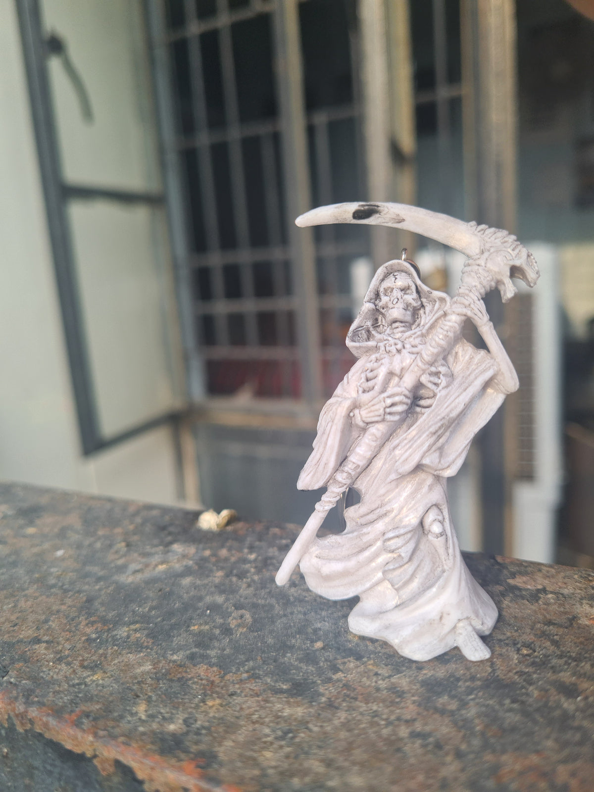 A small white skeleton figurine holding a scythe, mounted on a dark textured vertical surface with a blurred building background.